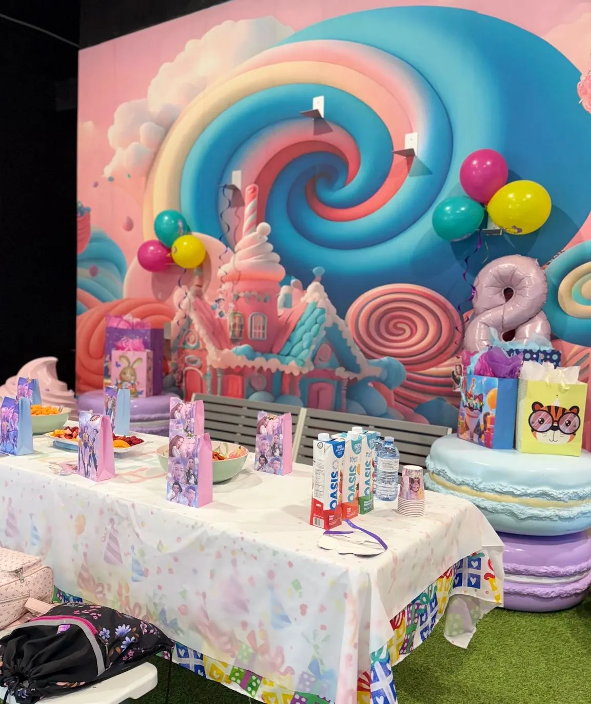 Table d'anniversaire garnie de sacs surprises, de fruits et de boissons devant la murale bonbon